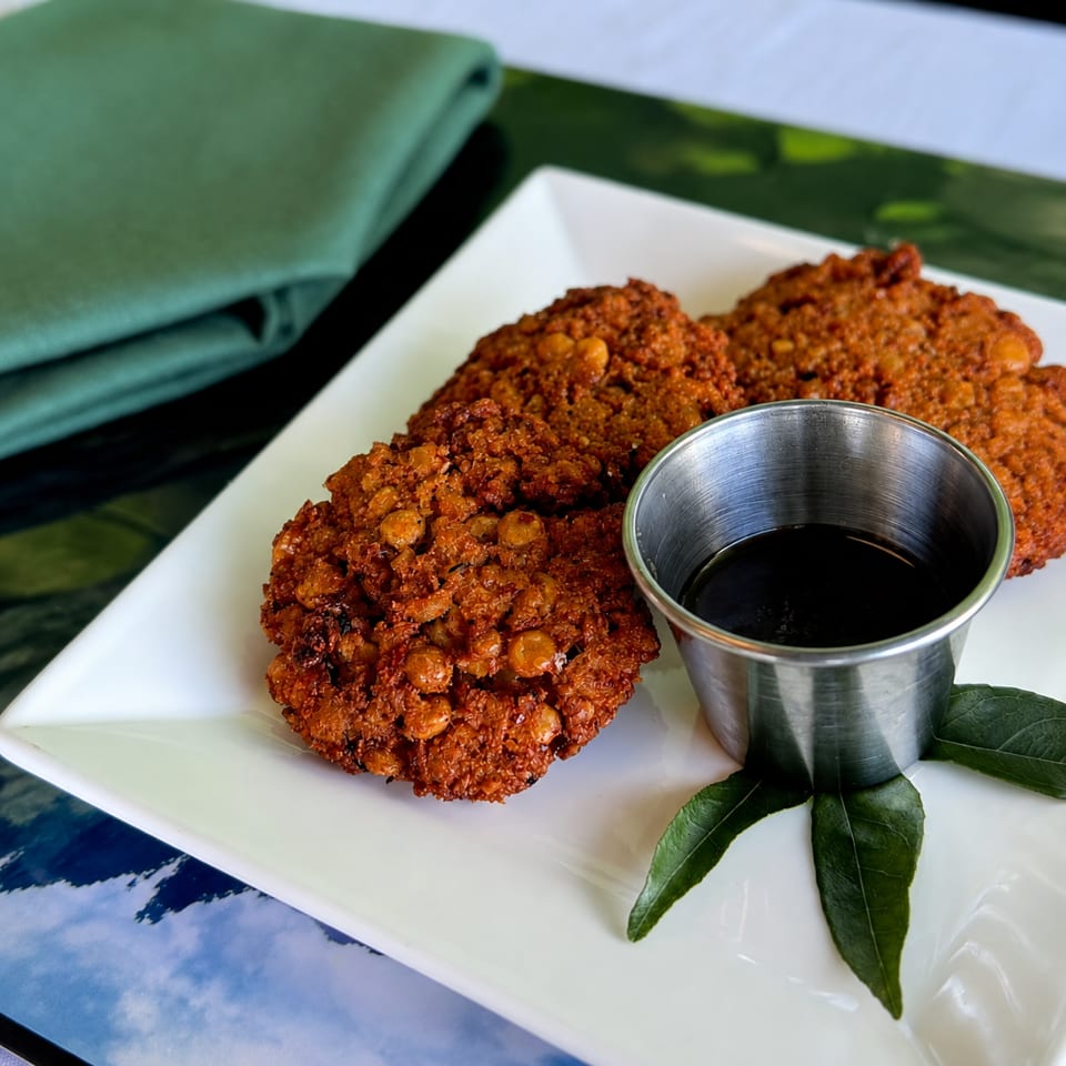 Crispy golden fritters studded with legumes sit on a white plate beside dark dipping sauce and green leaves.