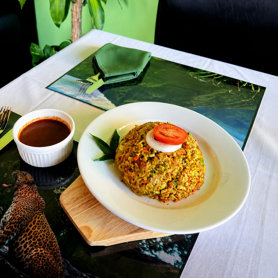 Spiced rice dish garnished with tomato and onion, served with dark sauce on a white plate at a tropical restaurant table.